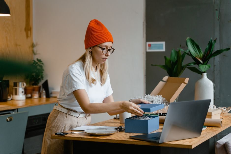 Woman packing a gift box with care at a cozy indoor workspace, using a laptop for guidance.